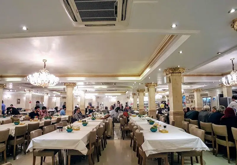 فضای داخلی لوکس و مدرن رستوران هانی مطهری با مشتریان در حال صرف غذا، میز و صندلی‌های چیده شده و لوسترهای زیبا.