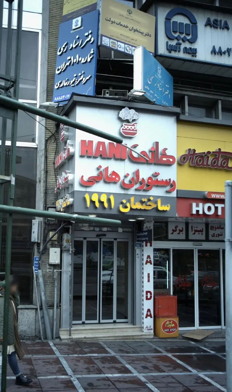 نمای بیرونی رستوران هانی مطهری در خیابان، با تابلوی مشخص رستوران هانی و سال تاسیس ۱۹۹۱. ورودی و پیاده‌رو مقابل رستوران دیده می‌شود.