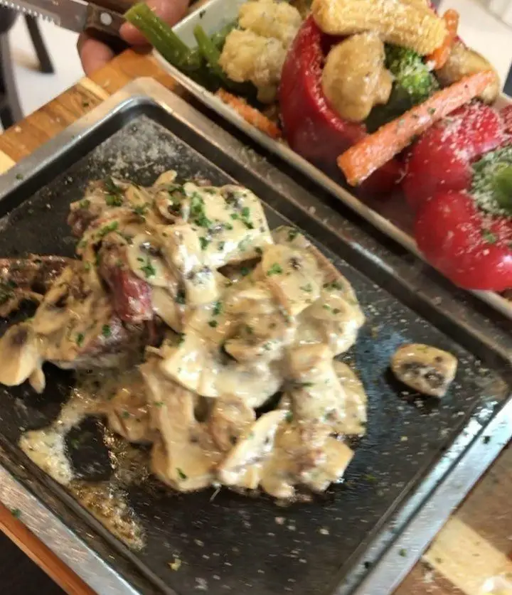 نمایی نزدیک از استیک یا فیله گوشت با سس قارچ خامه‌ای و سبزیجات تازه در رستوران هانی مطهری، تجربه‌ای لذت‌بخش از یک غذای خوشمزه.