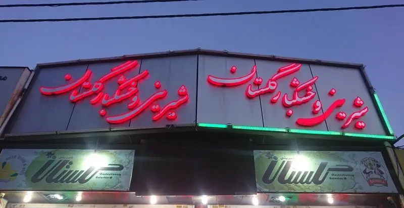 نمایی از تابلوی نئون قرمز شیرینی سرای گلستان (17) در شب با نورپردازی، شامل نوشته 'شیرینی خنک و تر گلستان' و تابلوی قنادی.