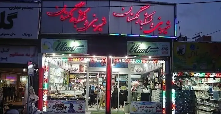 تصویر شب از نمای بیرونی شیرینی سرای گلستان، با تابلوی نئون درخشان، محصولات متنوع قنادی و دسر در ویترین‌های روشن فروشگاه