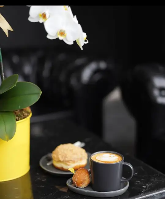 یک فنجان قهوه لاته با طرح روی فوم، دو دونات طلایی و یک گلدان ارکیده سفید زیبا روی میز در کافه لیژ. فضایی دلنشین برای مشتریان.