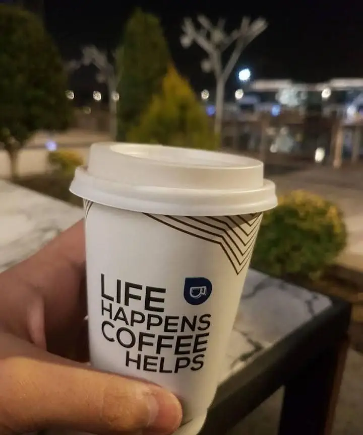 لیوان قهوه برند بالکافه با لوگو و شعار «زندگی اتفاق می‌افتد، قهوه کمک می‌کند» در دست مشتری، در فضای آرام و زیبای کافه در شب.