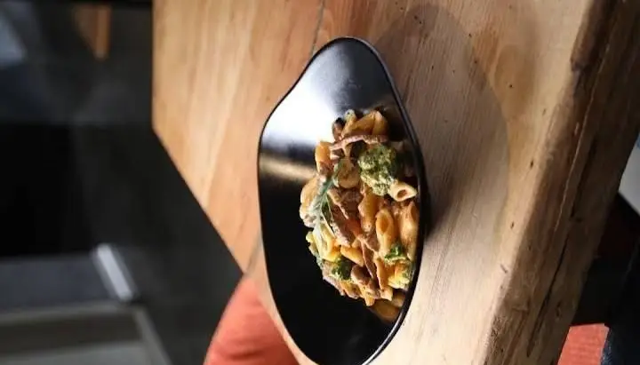 پاستای پنه کرمی با بروکلی تازه و تکه‌های مرغ در بشقاب مدرن مشکی، سرو شده روی میز چوبی، یکی از غذاهای محبوب بالکافه.