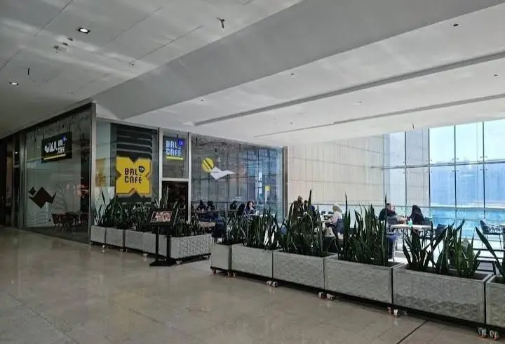 نمای بیرونی کافه بالکافه در یک مرکز خرید مدرن با مشتریانی در حال استراحت، فضای سبز و پنجره‌های بزرگ شیشه‌ای.