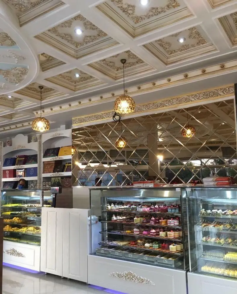 ویترین‌های پر از کیک و شیرینی‌های رنگارنگ در نان شیرینی ناز گل با سقف گچ‌بری شده و آینه‌کاری زیبا، محیط دلنشین قنادی.