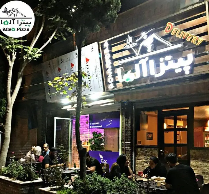 فضای بیرونی دنج پیتزا آلما در شب، با تابلوهای روشن و مشتریانی که از پیتزا و محیط لذت می‌برند. مناسب برای سفارش دلیوری.
