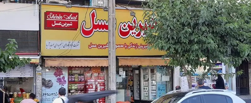تصویر واضح از نمای بیرونی فروشگاه شیرین عسل گیشا در تهران، با تابلوهای فارسی و انگلیسی. این شعبه محصولات غذایی و شیرینیجات شیرین عسل را ارائه می‌دهد.