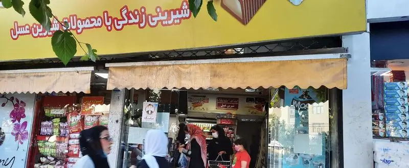 نمای بیرونی فروشگاه شیرین عسل گیشا در منطقه گیشا، با تابلوی زردرنگ و چشمگیر "شیرینی زندگی با محصولات شیرین عسل" و حضور مردم.