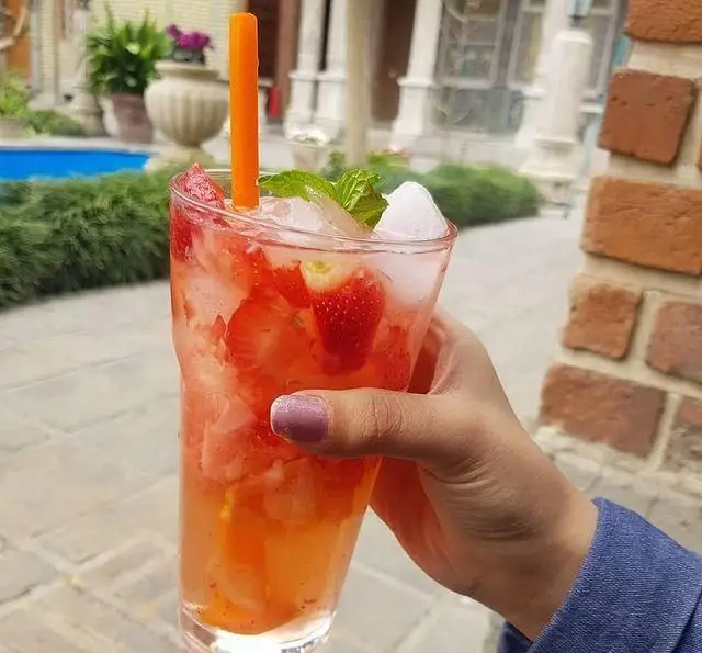 دست در حال گرفتن نوشیدنی خنک توت فرنگی و نعناع در کافه گرین، محوطه سرسبز موزه مقدم. لحظات دلنشین تابستانی در فضای باز.