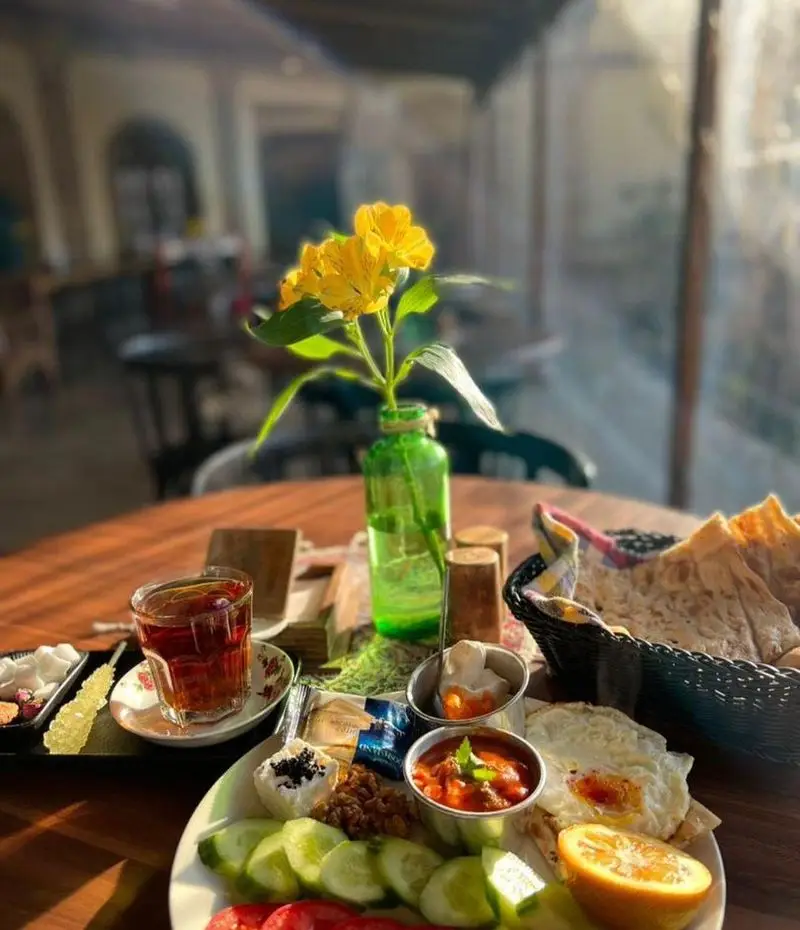 میز صبحانه ایرانی کامل و جذاب در کافه گرین (محوطه موزه مقدم) شامل نیمرو، نان، سبزیجات، چای و گل‌های زرد زیبا.