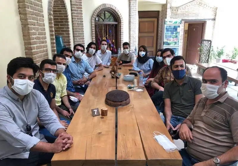 جمعی از افراد ماسک‌زده در کافه گرین موزه مقدم، دور میز چوبی بلند با کیک و نوشیدنی‌ها جمع شده‌اند.