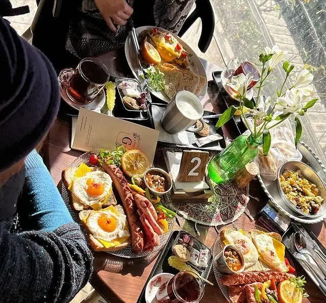 میز صبحانه و برانچ جذاب در کافه گرین (محوطه موزه مقدم) با انواع تخم مرغ، سوسیس، بیکن و نوشیدنی‌های گرم در فضایی دلنشین.