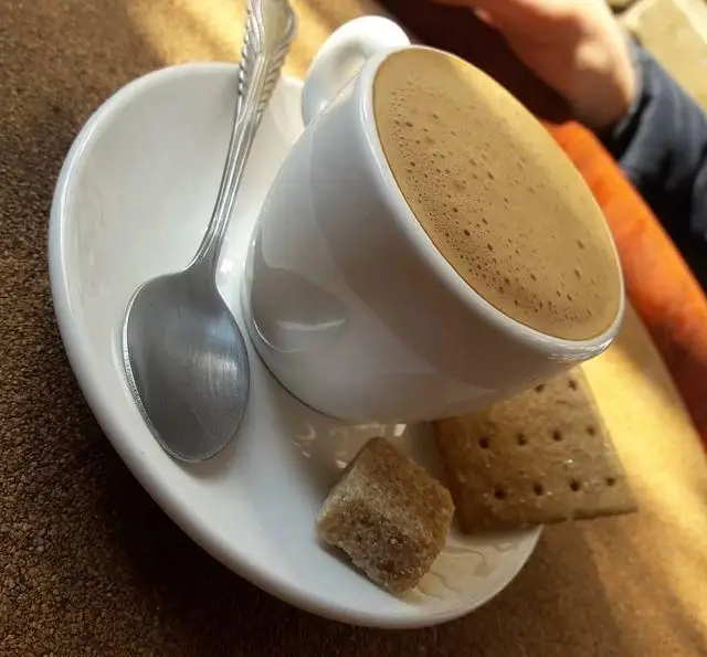 یک فنجان قهوه تازه با کف، قاشق، حبه قند قهوه‌ای و بیسکویت در کافه گرین (محوطه موزه مقدم)، لحظه‌ای دلنشین و آرامبخش.