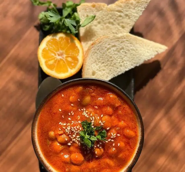 یک کاسه خوراک نخود داغ و خوشرنگ کافه گرین (موزه مقدم) با نان تازه، نصف لیمو و سبزیجات، تزیین شده با کنجد و جعفری.