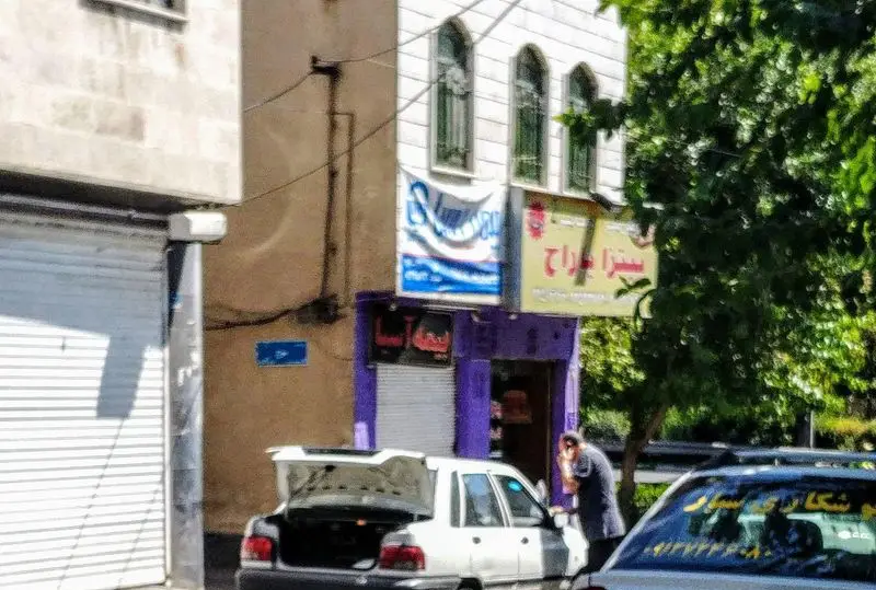 نمایی آفتابی از بیرون پیتزا پاراج، شامل تابلوی زرد رنگ، ورودی بنفش مغازه و یک خودروی سفید با صندوق عقب باز در جلوی آن.