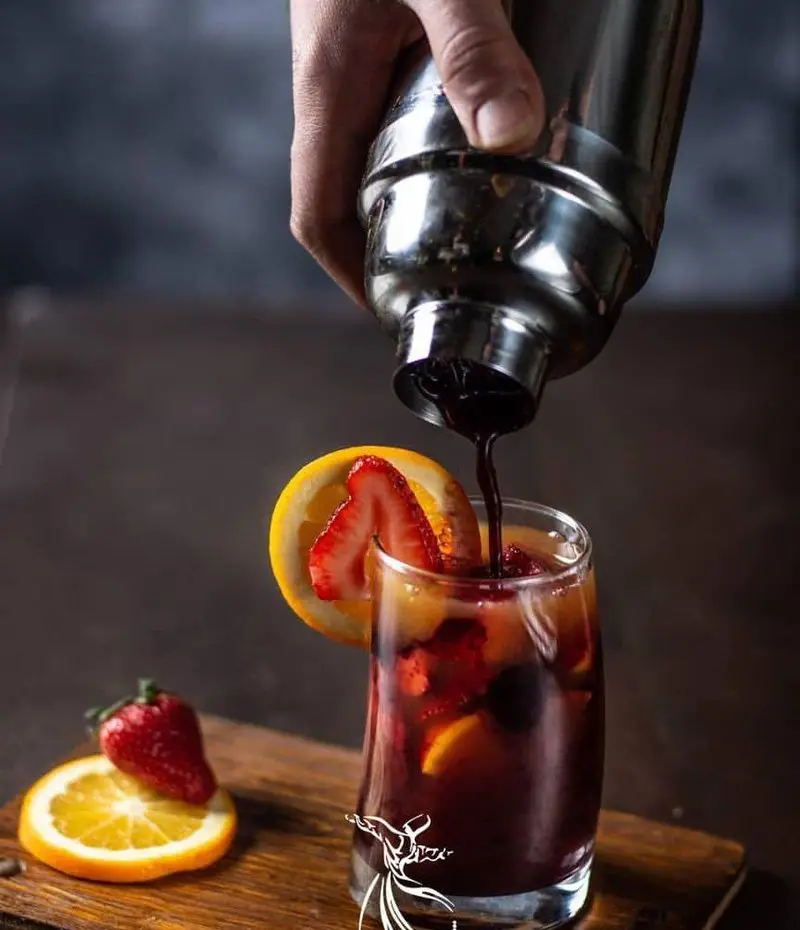 بارتندر کافه رستوران درویش در حال ریختن کوکتل میوه‌ای تازه و خنک از شیکر به لیوان، با تزیین پرتقال و توت‌فرنگی.