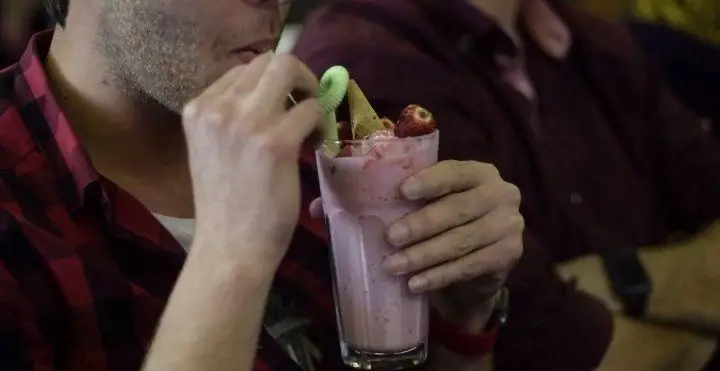 تصویر مشتری در کافه رستوران درویش که با نی سبز، میلک شیک توت فرنگی تازه و خنک خود را با تزئین بستنی و توت فرنگی میل می‌کند.