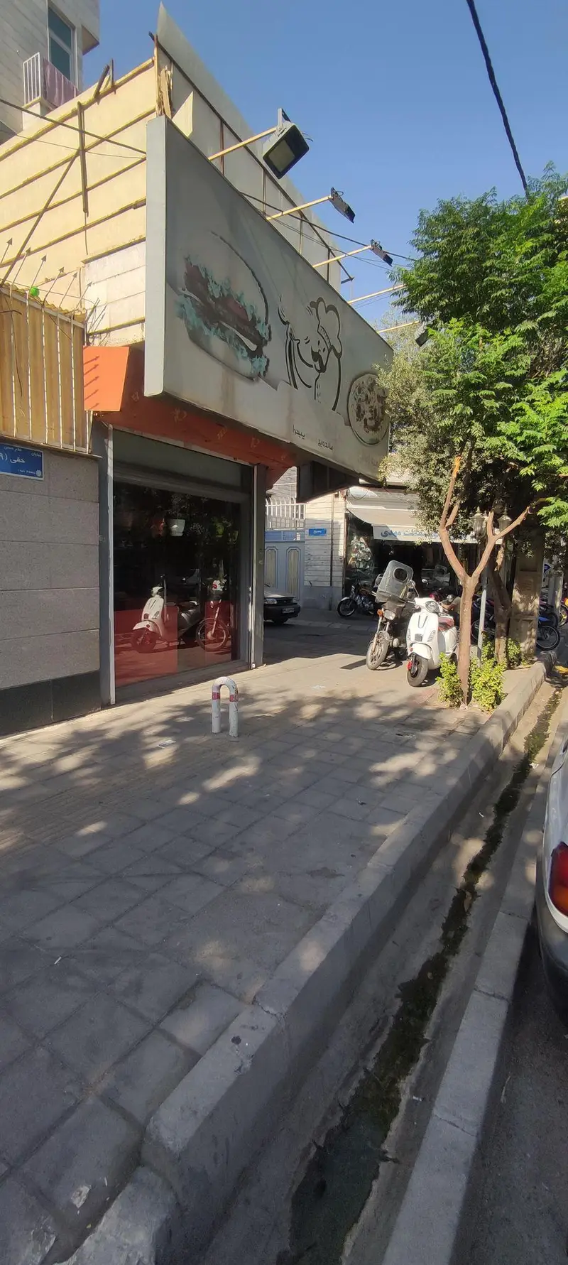 نمای خارجی ساندویچی دولپی با تابلوی جذاب حاوی عکس برگر و یک سرآشپز. اسکوترهای سفید و قرمز در ویترین، پیاده‌روی سنگفرش و موتورها در خیابان.