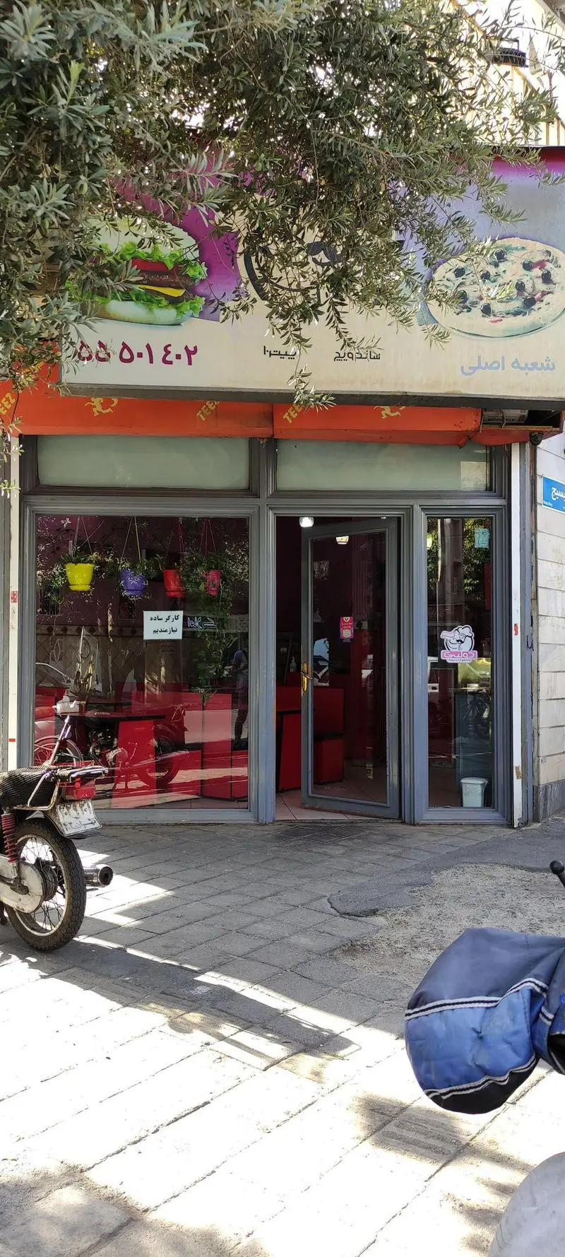 نمای بیرونی رستوران ساندویچی دولپی، شامل تابلوی بزرگ با تصویر برگر و پیتزا. شماره تماس، درخت و موتور در جلوی درب.