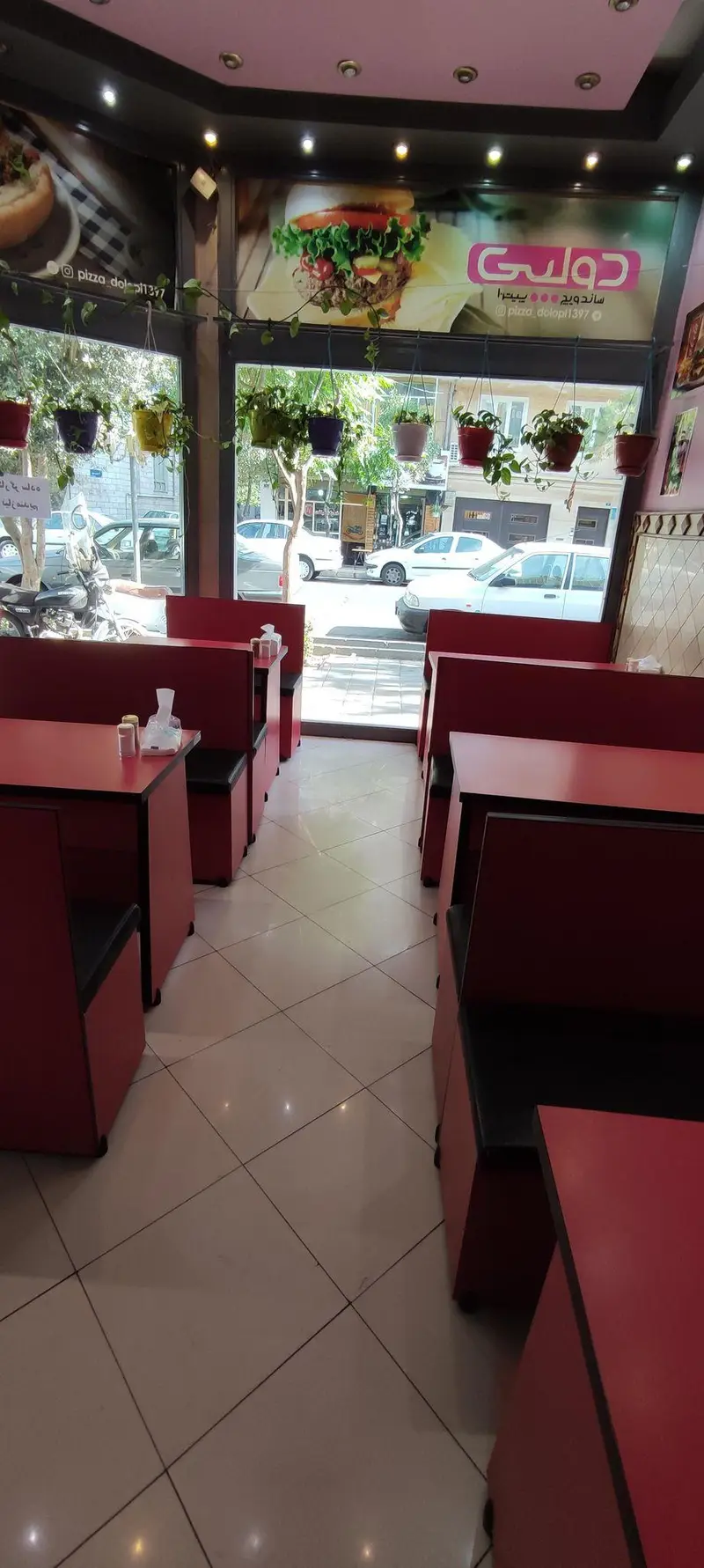 فضای داخلی ساندویچی دولپی با میز و صندلی‌های قرمز، دکوراسیون فست فود و نمای خیابان. دارای بنرهای همبرگر، پیتزا و آدرس Dolapi.