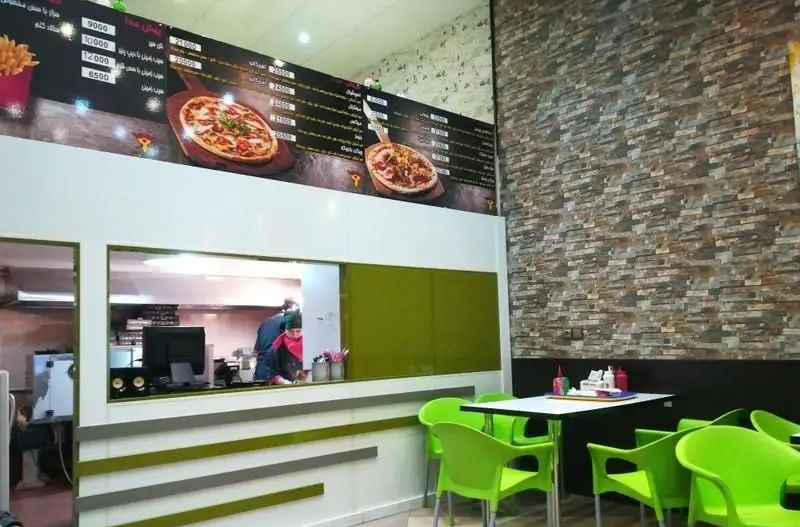 فضای داخلی پیتزایی پیتزا چیمن با دکوراسیون مدرن، میز و صندلی‌های سبز، منوی بالای سر و آشپزخانه باز برای سرو پیتزا و سیب‌زمینی.