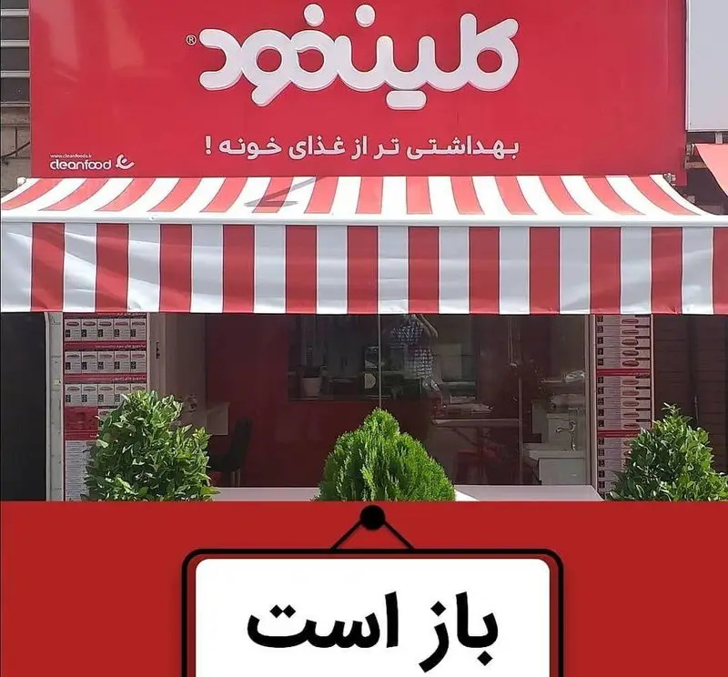 تصویر نمای خارجی فست فود کلین فود با تابلو قرمز رنگ، سایبان راه راه قرمز و سفید و اعلامیه 'باز است'، نشانگر خدمات غذای بهداشتی.