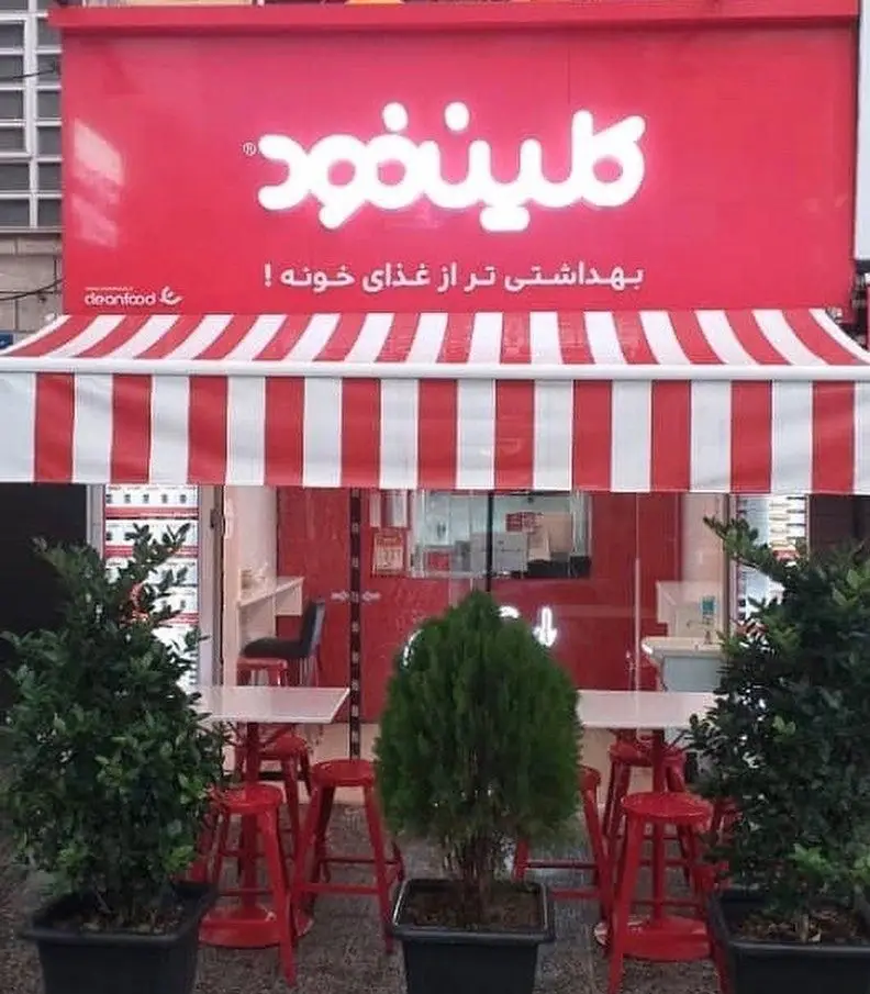 تصویر نمای خارجی شعبه کلین فود با دکوراسیون قرمز و سفید، فضای باز، تابلو روشن و شعار بهداشتی تر از غذای خونه.