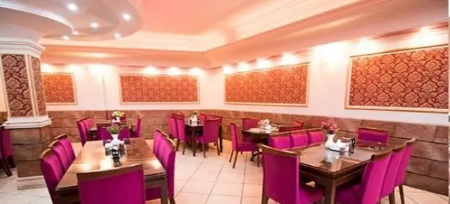رستوران ایرانی کلانتر: فضای داخلی زیبا با صندلی‌های صورتی لوکس، میزهای چوبی، دیوارپوش‌های طرح‌دار و نورپردازی دلپذیر آماده پذیرایی.
