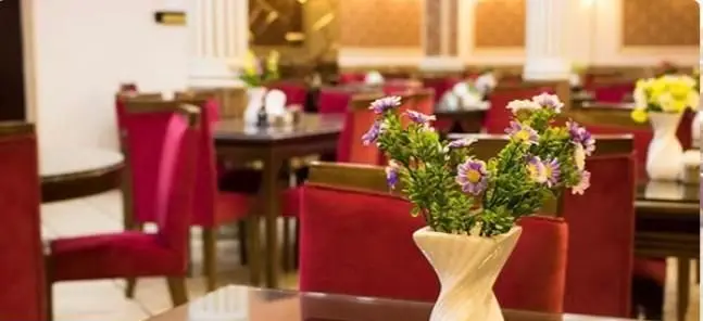 نمای داخلی شیک رستوران ایرانی کلانتر با میزهای چوبی تیره، صندلی‌های قرمز راحت و گلدان‌های گل، فضایی دلنشین برای میزبانی.