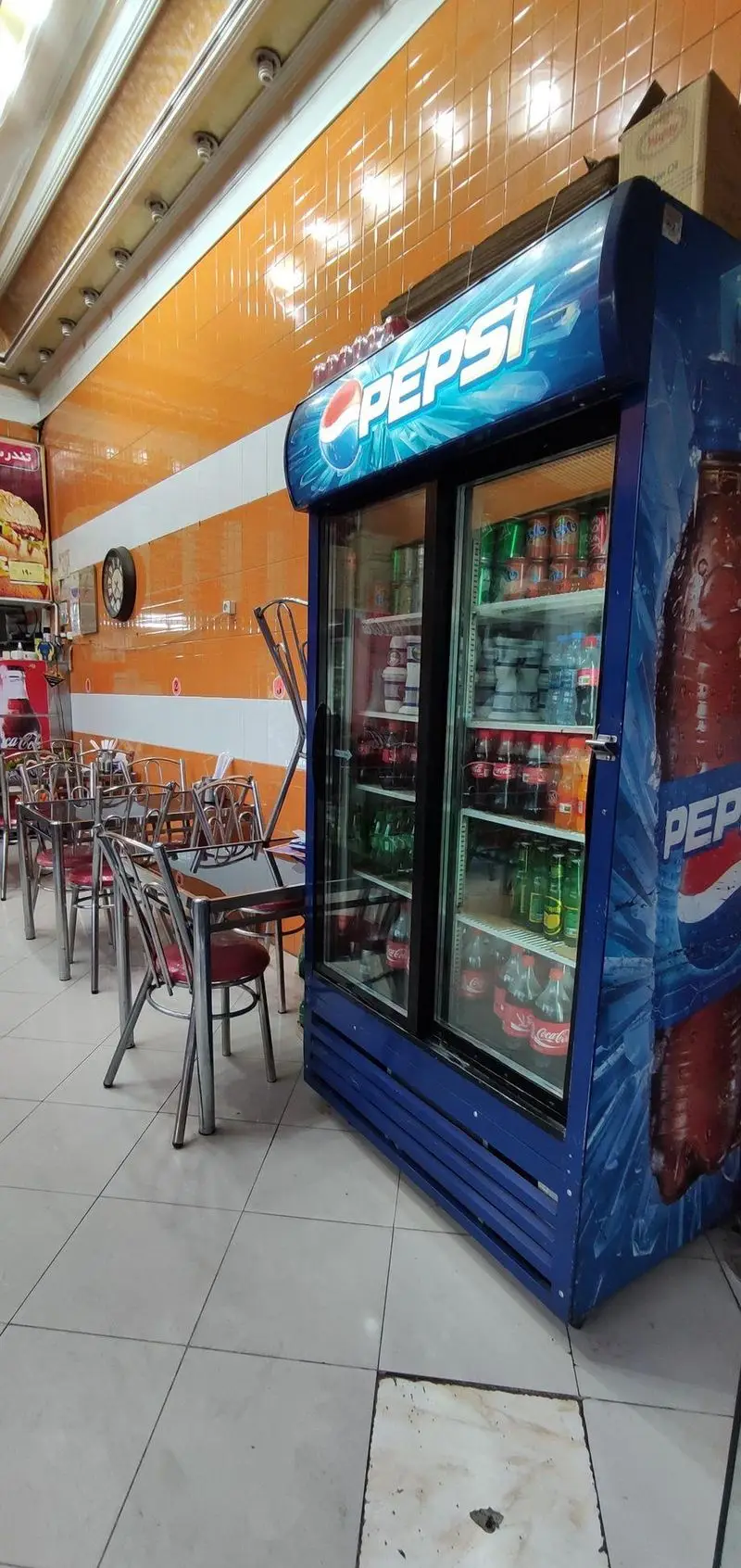 فضای داخلی رستوران هایس برگر با میز و صندلی‌های مدرن، یخچال نوشیدنی‌های پپسی و کوکاکولا، و دیوار نارنجی کاشی‌کاری شده.
