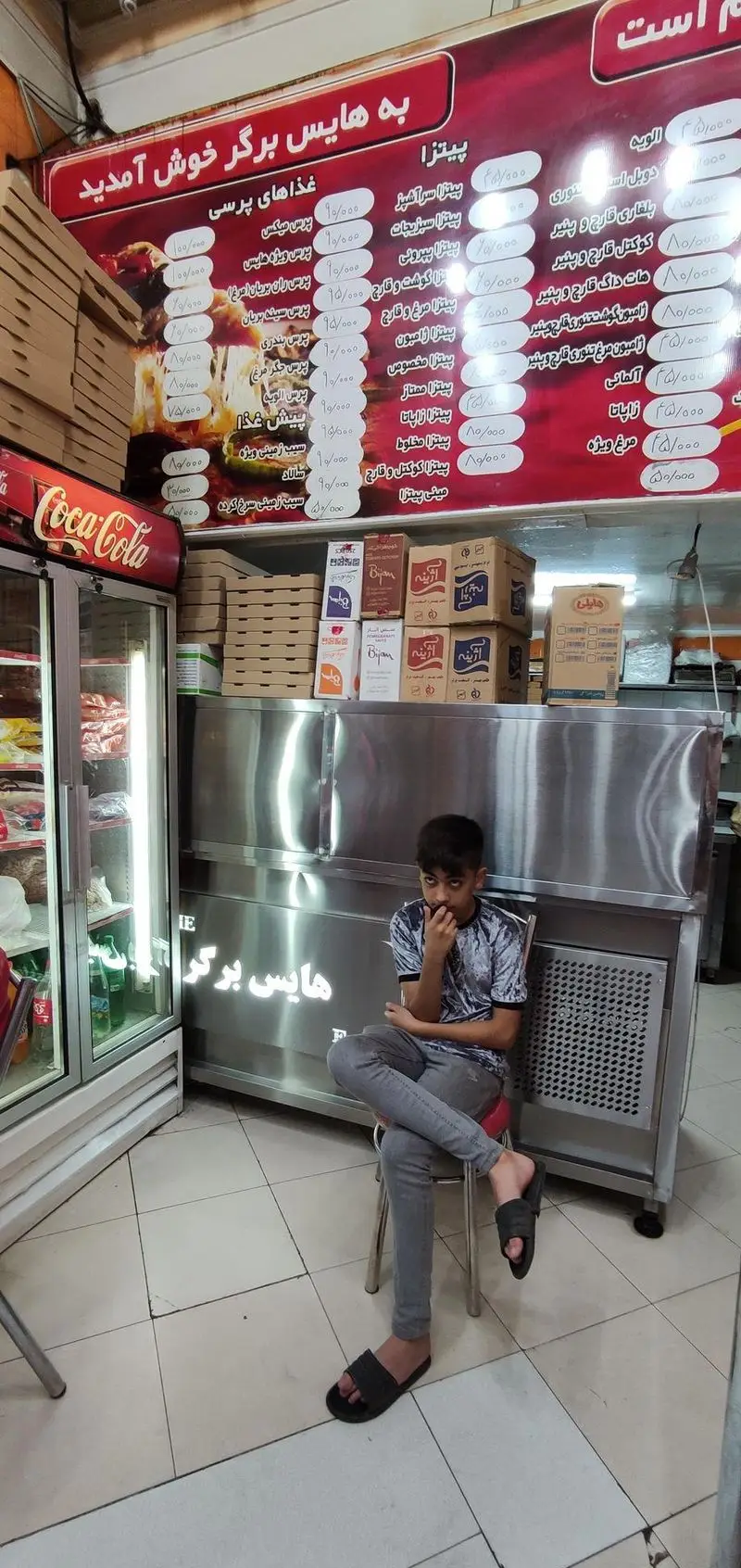 هایس برگر: نمای داخلی رستوران فست فود با منوی پیتزا و برگر، یخچال کوکاکولا، جعبه‌های غذا و پسربچه‌ای نشسته کنار پیشخوان.