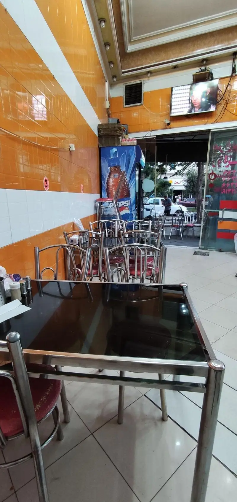 نمایی داخلی از رستوران هایس برگر با چیدمان میز و صندلی‌های استیل، یخچال پپسی، تلویزیون و دیوارهای نارنجی، محیطی مرتب برای فست فود.