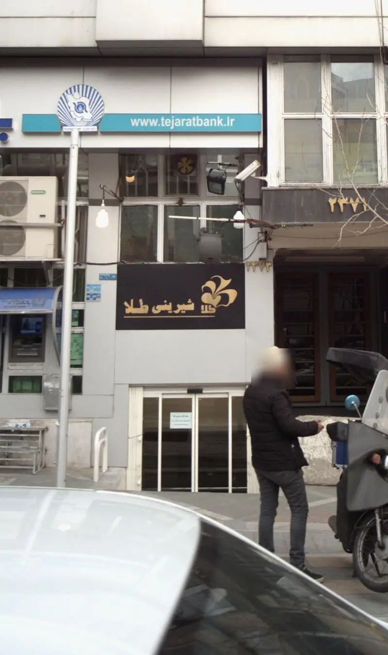 عکس نمای بیرونی قنادی "شیرینی طلا" با ورودی اصلی، تابلوی مشکی و لوگوی طلایی، در کنار تابلوی بانک تجارت در تهران.