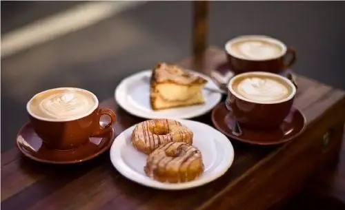 سه فنجان قهوه لاته با طرح، یک تکه کیک و دو دونات خوشمزه روی میز چوبی در کافه نان دکتر کافه، مناسب دورهمی عصرانه.