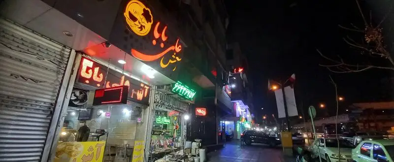آش علی بابا: نمای شبانه یک رستوران ایرانی درخشان با تابلوی نئون و لوگوی متمایز در یک خیابان شهری پرتردد.
