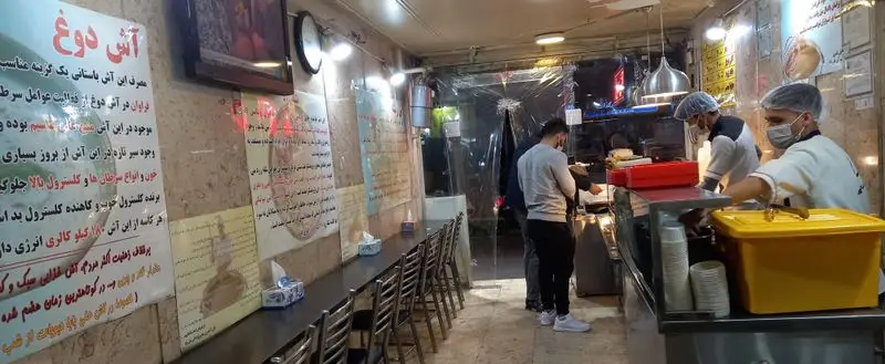 تصویری از داخل آش علی بابا؛ آشپزان با ماسک و کلاه آشپزی مشغول آماده‌سازی آش دوغ و خوراکی‌های دیگر برای مشتریان هستند.