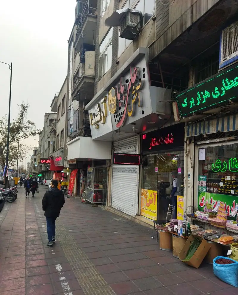 تصویری از نمای خیابان شلوغ شهری با مغازه آش علی بابا، عطاری هزار برگ و سایر کسب و کارهای محلی در تهران، مردم در حال عبور و مرور