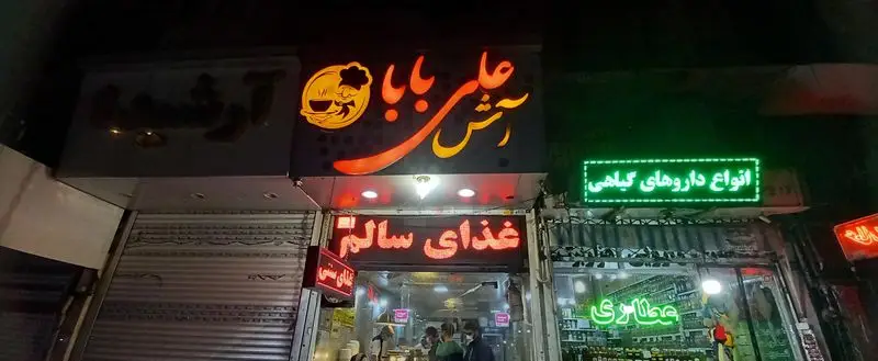 تابلو آش علی بابا با لوگوی سرآشپز، رستوران غذای سالم و سنتی. در کنار آن، عطاری با انواع داروهای گیاهی و محصولات طبیعی.