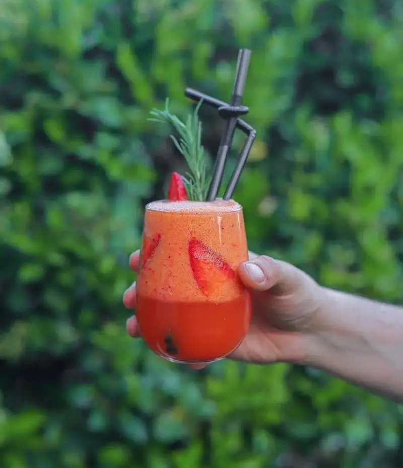 نوشیدنی اسموتی لایه لایه توت فرنگی و رزماری تازه کافه بینج، با نی‌های جذاب در دست در فضای سبز و دلنشین طبیعت