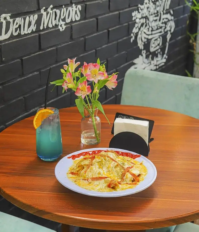 میز چوبی زیبای کافه بینج با غذای کرمی خوشمزه، نوشیدنی آبی خنک با پرتقال، گل‌های صورتی و دیوار آجری مشکی شیک.