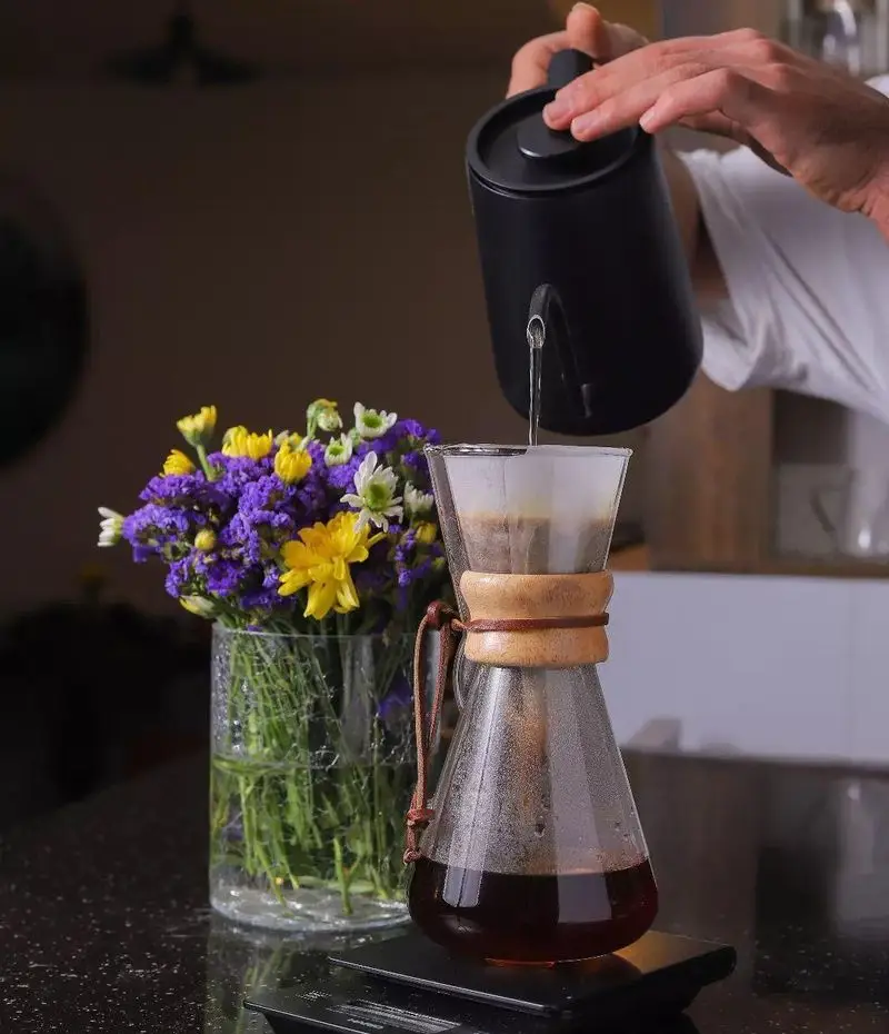 کافه بینج: تهیه قهوه دمی با کمکس توسط باریستا. آب‌ریزی دقیق با کتری مشکی روی قهوه. گلدان گل‌های زیبا در کنار. تجربه نوشیدن قهوه باکیفیت و تازه‌دم در کافه.