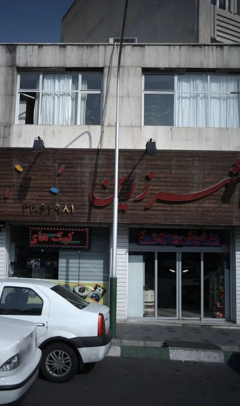 شیرینی‌فروشی شهر زرین: نمای بیرونی یک قنادی در خیابان، با تابلوی روشن، ویترین جذاب و خودروهای پارک شده در جلو.