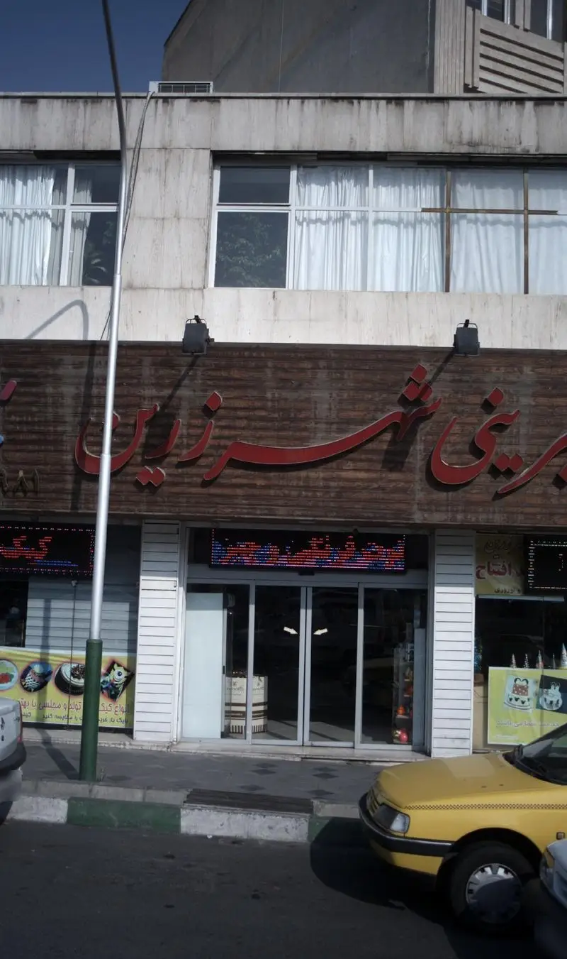 شیرینی شهر زرین: نمای بیرونی قنادی محبوب با سردر چوبی، تابلوی فارسی قرمز و ورودی شیشه‌ای. تاکسی زرد در خیابان تهران.