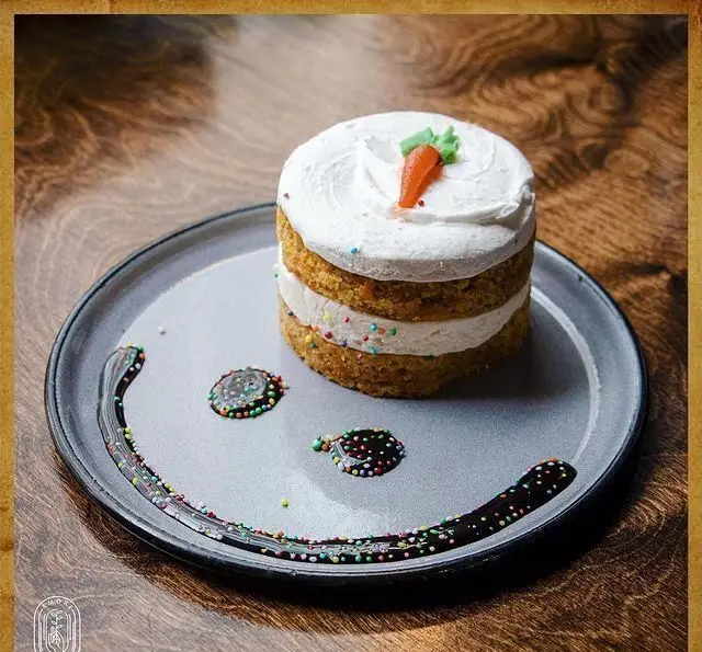 یک کیک هویج مینی جذاب و خوشمزه از کافه آموره با لایه‌های خامه سفید، تزئین اسپرینکل و سس شکلات فانتزی روی میز چوبی