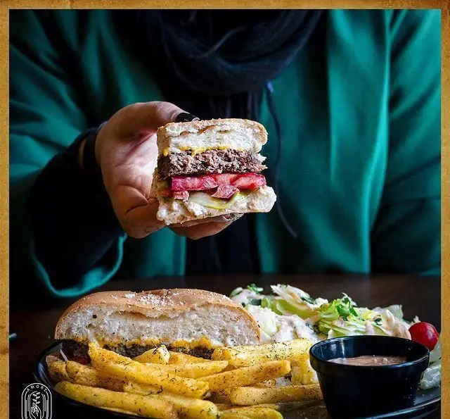 تجربه یک برگر لذیذ با سیب‌زمینی‌های ترد و سالاد تازه در کافه آموره، آماده سرو برای شما. نیمی از برگر در دست.