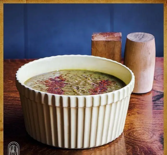 تصویری زیبا از کاسه بزرگ آش سبز یا عدسی گرم و سنتی، تزئین شده با ادویه قرمز، روی میز چوبی در فضای دلنشین کافه آموره.