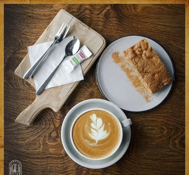 یک فنجان لاته آرت زیبا و یک برش کیک لذیذ روی میز چوبی در کافه آموره، انتخابی عالی برای صبحانه یا عصرانه دلپذیر.
