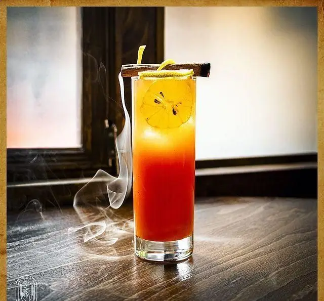 تصویری زیبا از کوکتل دودی و خوش‌رنگ کافه آموره با گارنیش لیمو و چوب دارچین روی میز چوبی، تجربه‌ای دلچسب.