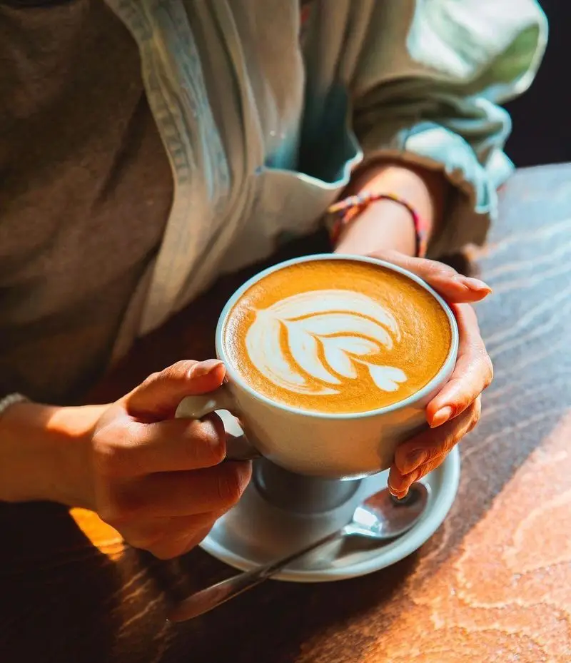 فنجان لاته داغ با لاته آرت هنرمندانه در دستان مشتری کافه آموره. لذت نوشیدن قهوه با کیفیت در محیطی آرام و دلنشین.
